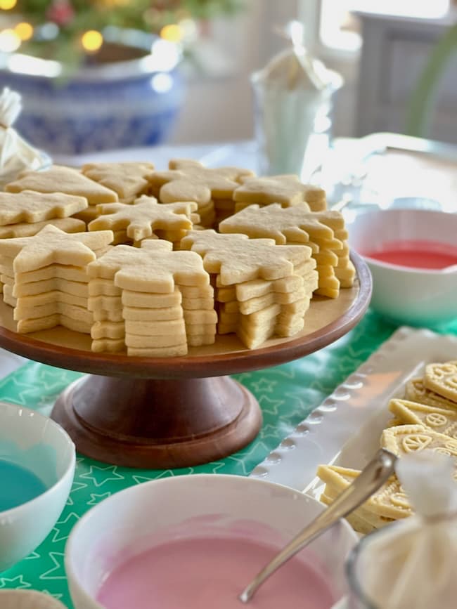 A wooden stand with stacks of star and tree-shaped cookies. Nearby, bowls filled with pastel-colored icing in pink, blue, and red are ready for decorating, and more cookies in various shapes are on a tray.