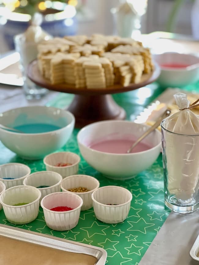 A table set for cookie decorating, featuring a plate of cookies, bowls of colorful icing in pink and blue, and small cups filled with various sprinkles and decorations. A piping bag is visible next to the setup.