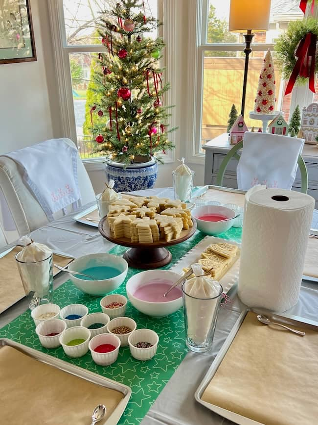 A festive table setup for cookie decorating, featuring a centerpiece of cookies on a stand, bowls of colorful icing, and sprinkles. The background includes a decorated Christmas tree and holiday-themed decor by the window.