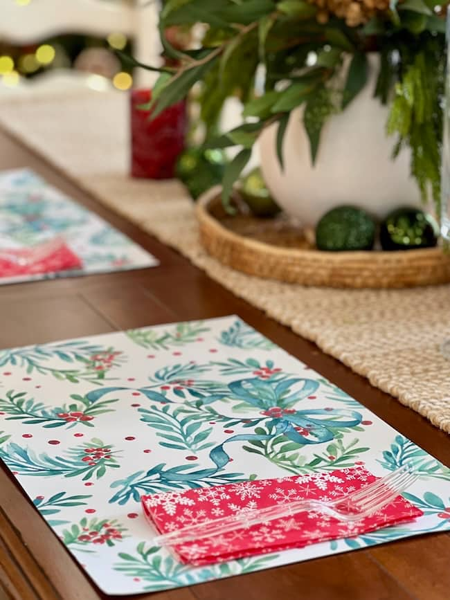 Festive holiday table setting with colorful placemats featuring floral and leaf patterns. A decorative arrangement with greenery and ornaments is in the background on a woven mat. A red napkin with a snowflake design holds cutlery.