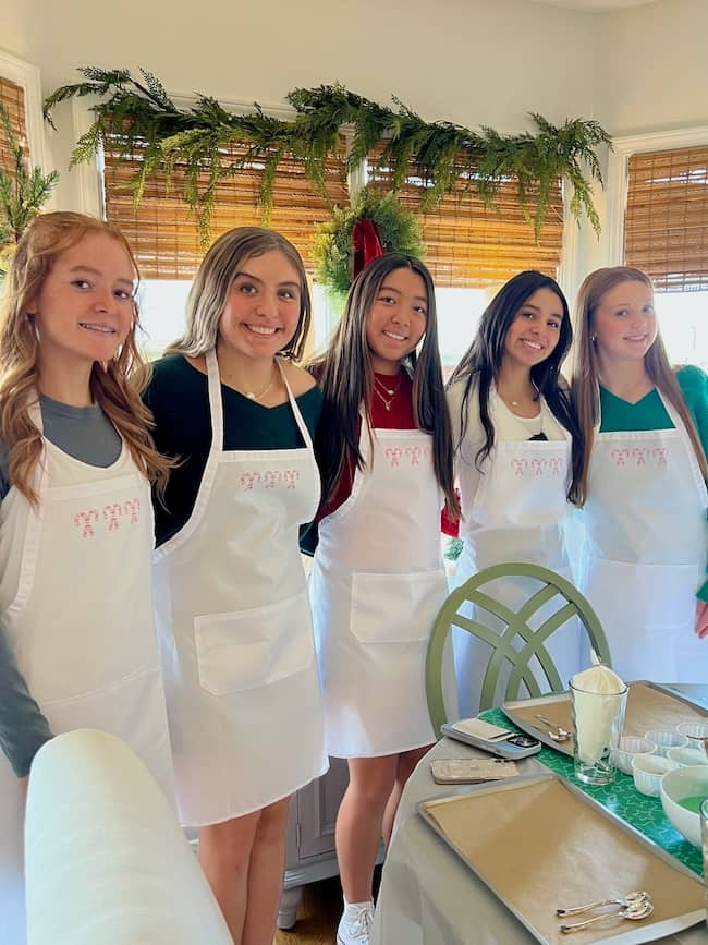 Five young women wearing white aprons stand together in a festive dining room. The table is set with plates, napkins, and green decorations. Greenery and red bows adorn the windows in the background. They are smiling at the camera.