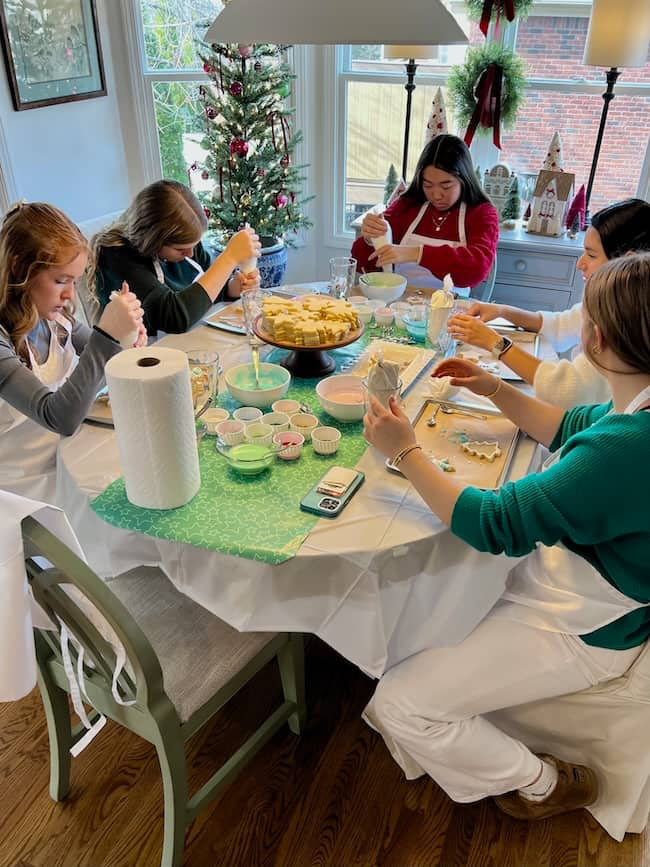 Five people seated around a table are decorating cookies with icing. The table is covered with bowls of different-colored icing, cookie cutters, and paper towels. A small Christmas tree and festive decorations are visible in the background.