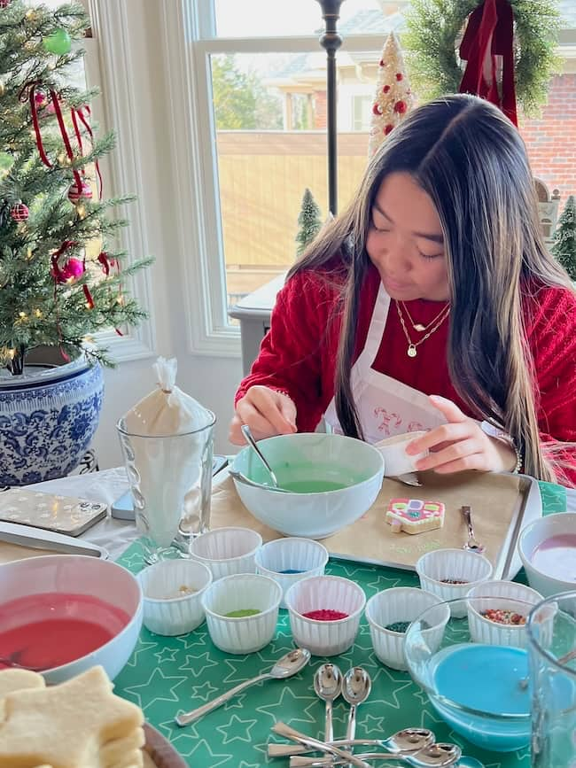 A woman decorates cookies at a table covered with bowls of colored icing and sprinkles. She wears a red sweater and white apron. A decorated Christmas tree and small pine trees are in the background, near a window.