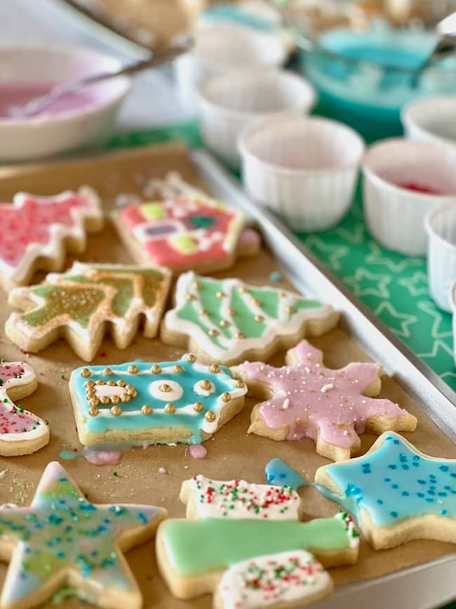 A tray of colorful, decorated sugar cookies in various shapes, including stars, trees, and snowflakes. Bowls of icing and sprinkles are visible in the background, suggesting a festive cookie decorating session.