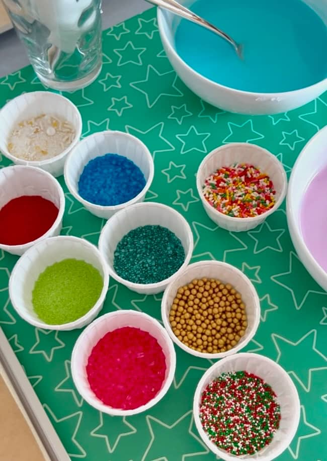 A variety of colorful sprinkles in small white containers are arranged on a green mat with star patterns. There are also two bowls containing blue and pink icing in the background.