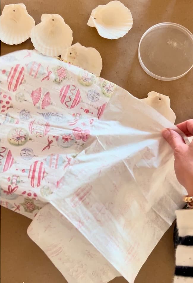 A person is carefully placing a decorative napkin with colorful Easter egg patterns onto a table. Nearby are white seashells, perfect for crafting unique holiday ornaments, and a small clear container. The person is wearing a striped sleeve, adding to the festive creativity of this decoupage project.