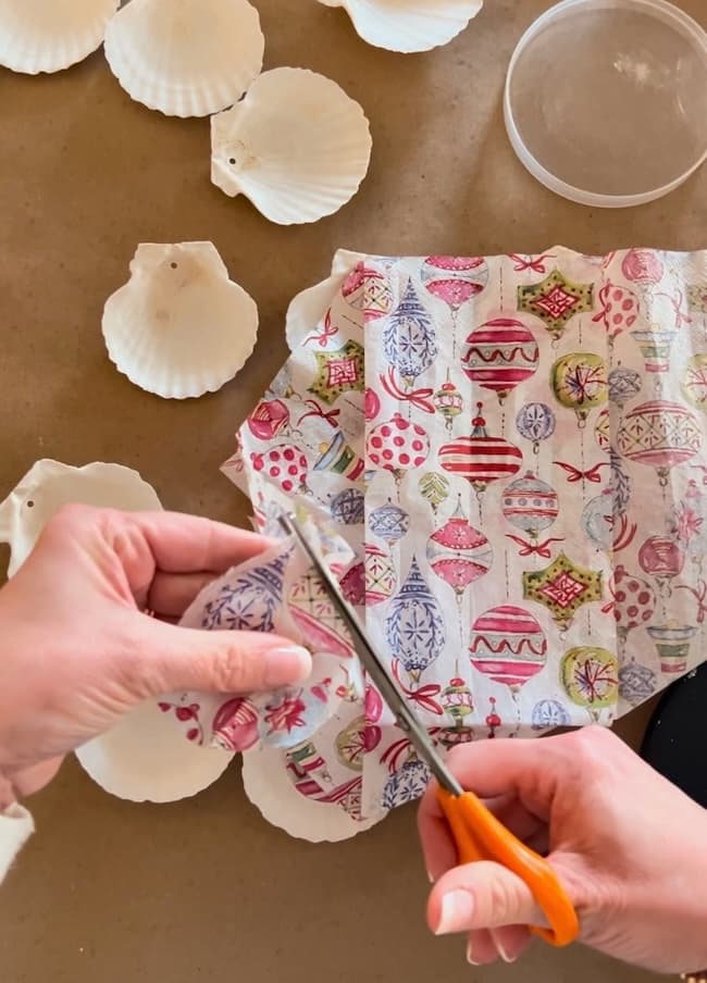 Hands using scissors to cut festive fabric adorned with holiday ornaments. Several white seashells, perfect for creating unique Seashell Christmas Ornaments, are scattered on the table, ready for a creative holiday craft project.