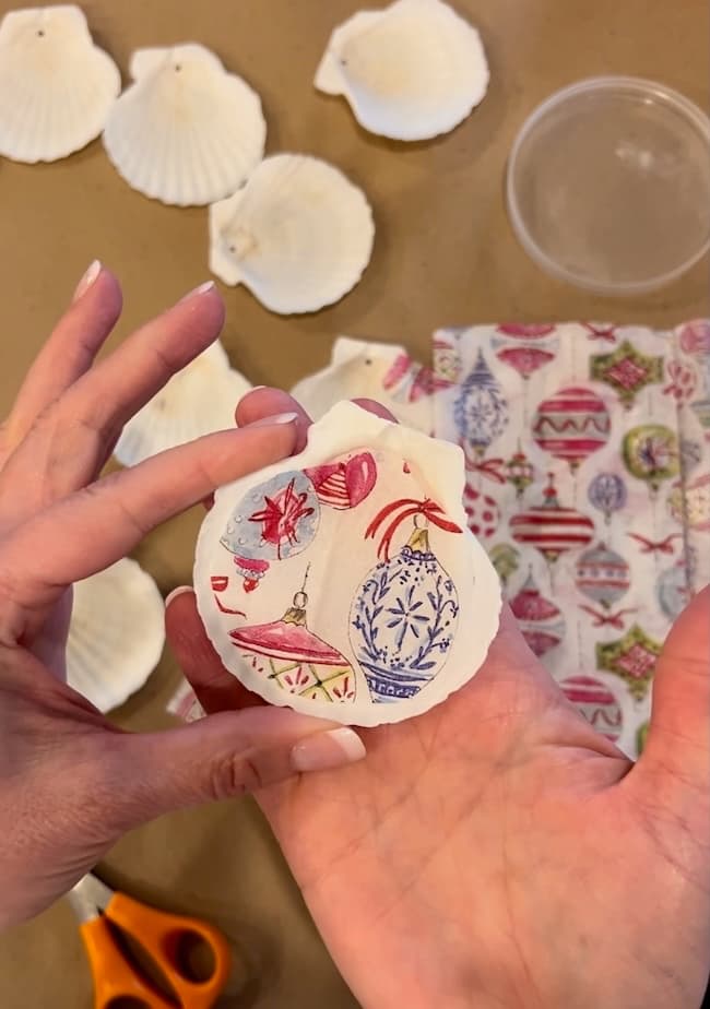 A person holding a seashell Christmas ornament adorned with colorful holiday designs. Other seashells, along with scissors and patterned paper for decoupage, are scattered on the table, enhancing the festive holiday craft scene.