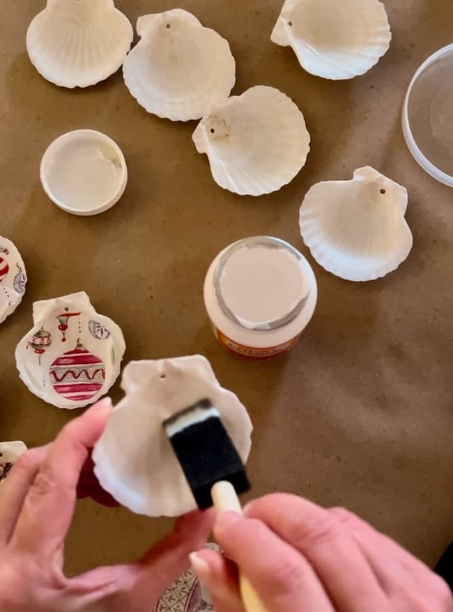 Hands are meticulously painting seashells with intricate designs using a brush. Some shells boast red and black patterns, perfect as Decoupage Seashell Christmas Ornaments, while others remain blank. Nearby, a bottle of paint or glue sits on the brown surface, ideal for this Holiday Craft activity.