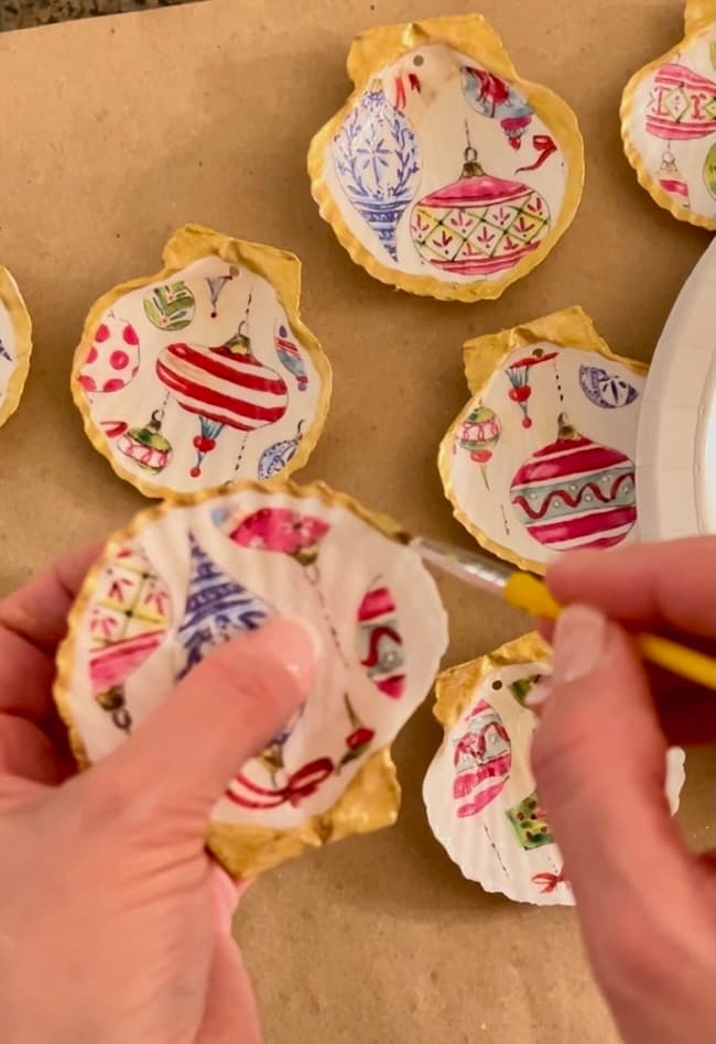 Hands decorating scallop shells with festive patterns, transforming them into seashell Christmas ornaments. The beautifully adorned shells rest on a brown surface, showcasing a delightful holiday craft.