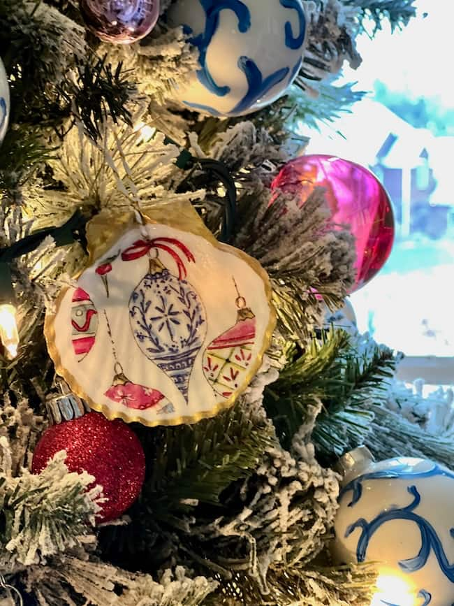 A close-up of a Christmas tree adorned with colorful ornaments, including a decorated white ornament with festive decoupage designs, pink and red round ornaments, and snow-dusted branches, set against a softly lit background.