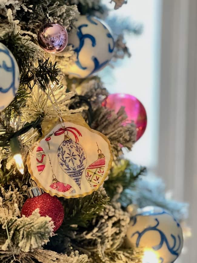 Close-up of a decorated Christmas tree with a variety of ornaments, including a painted round ornament with blue patterns and colorful baubles. The tree, showcasing holiday craft elements like DIY decoupage pieces, is dusted with artificial snow and softly lit, creating a festive atmosphere.