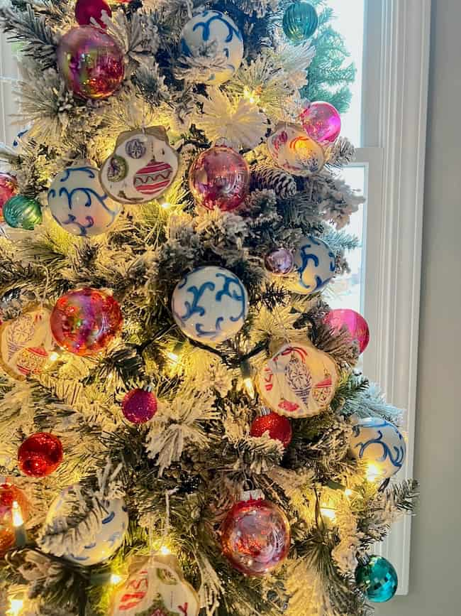 A festive Christmas tree adorned with colorful ornaments, including handmade seashell Christmas ornaments, is illuminated by warm string lights. The tree's branches are dusted with faux snow, and a window is visible in the background, completing this charming holiday craft scene.