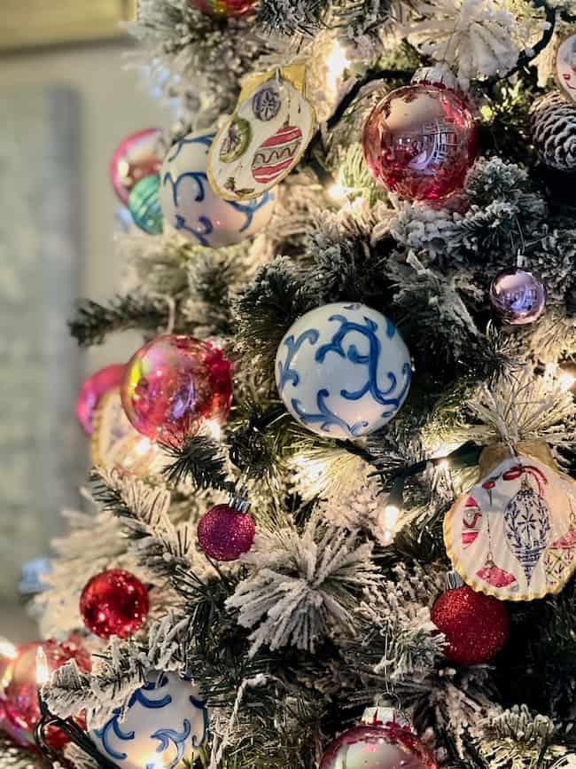 A close-up of a decorated Christmas tree with snow-dusted branches. Among the ornaments are large shiny pink balls, intricate blue and white patterned globes, colorful egg-shaped decorations, and handmade seashell Christmas ornaments, all lit by warm lights.