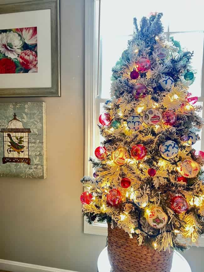 A small Christmas tree adorned with red and white ornaments and lights, including handcrafted seashell Christmas ornaments, sits in a woven basket. On the wall behind, two framed artworks feature floral and bird designs. The scene is illuminated by natural light from a window.