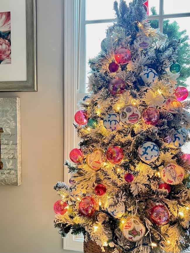 A Christmas tree adorned with pink and blue ornaments, including DIY decoupage creations, is lit with white string lights. It stands next to a window, allowing natural light to filter in. The surrounding walls display framed artwork.