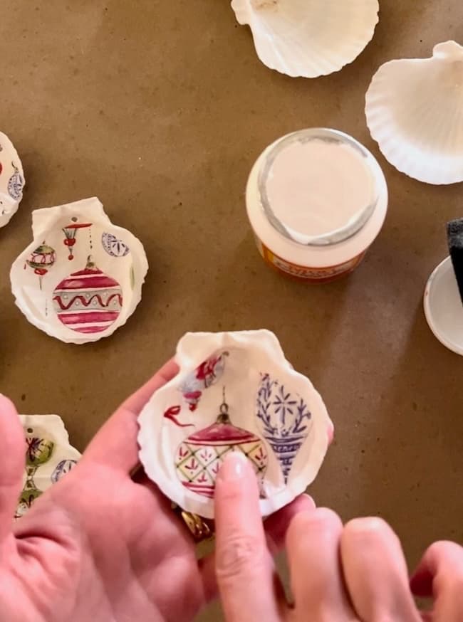 A person holds a seashell painted with colorful Christmas ornaments, showcasing their delightful holiday craft. Several other painted shells are scattered on the table. Nearby, there's an open jar of white paint or glue, perfect for decoupage creations.