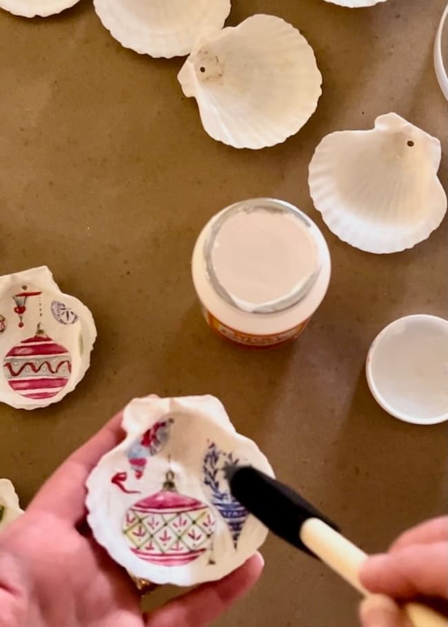 Hands holding a seashell painted with colorful ornaments, a brush delicately applying glaze. This holiday craft scene has several other white seashells and a jar of glue resting on the brown surface, perfect for creating unique decoupage or even Seashell Christmas Ornaments.