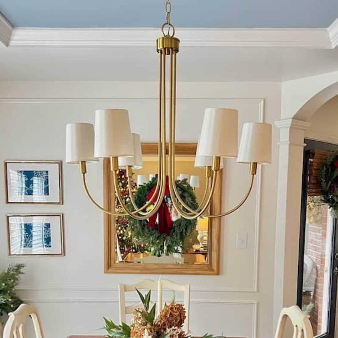 In the dining room, a brass chandelier with white lampshades elegantly illuminates the table adorned with floral decorations. A wreath with a red bow is reflected in the mirror, complementing framed pictures on the wall, offering charming breakfast nook lighting ideas.