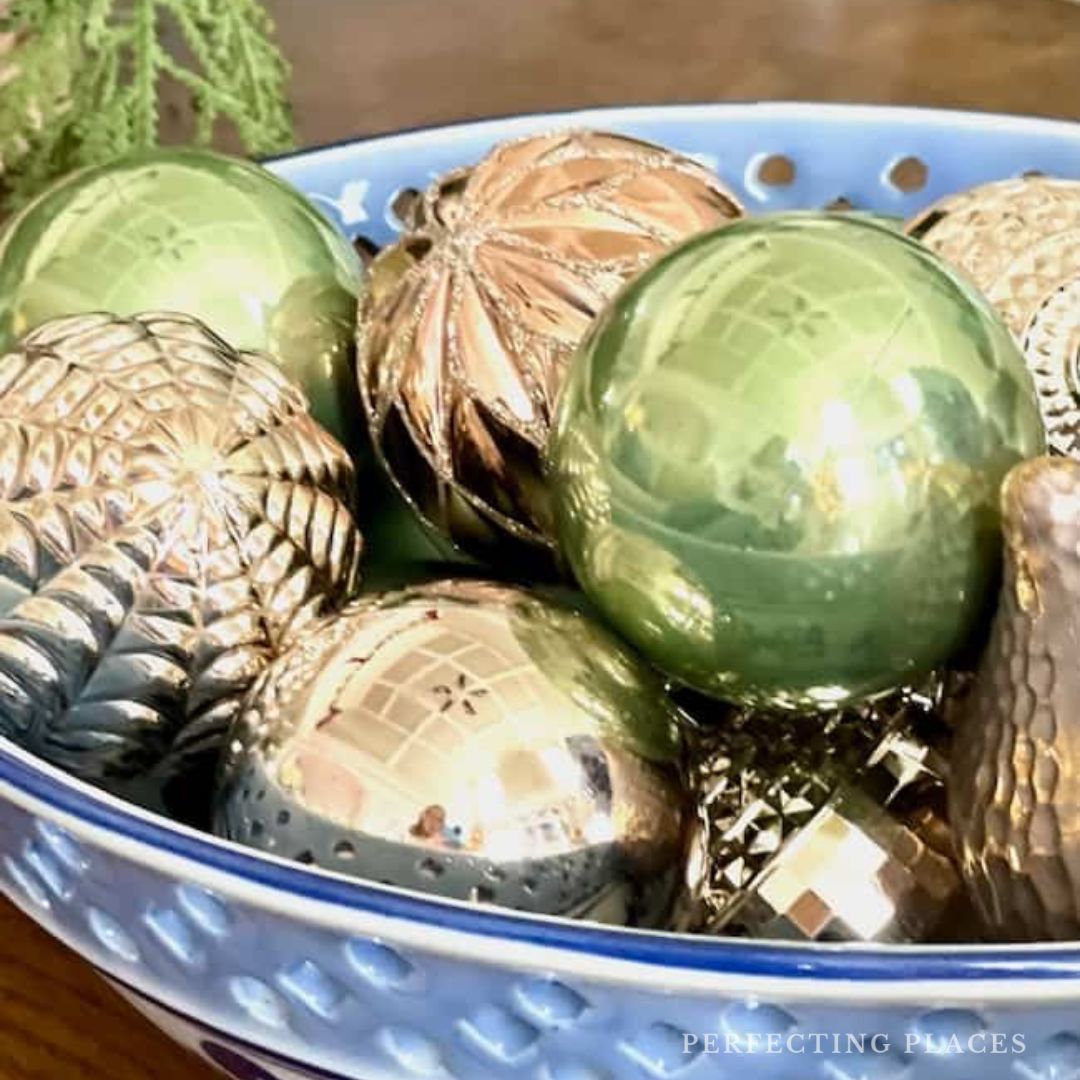 On a wooden surface sits a blue ceramic bowl, dazzling with shiny green and gold Christmas ornaments. Smooth spheres and textured pine cones capture the essence of December 2024, evoking the magic of Seven on Saturday festivities.