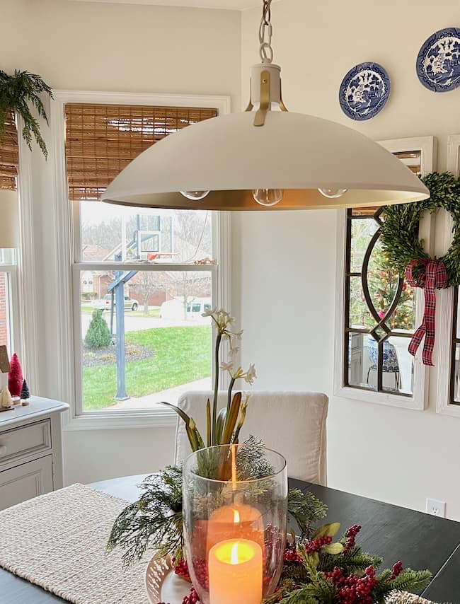 In a cozy dining room, Style Inspiration comes to life with Dining Room Lighting featuring a large pendant light above. A lit candle in a glass holder graces the table adorned with greenery, while windows with bamboo shades reveal a backyard basketball hoop. Holiday wreaths and decorative plates embellish the walls.