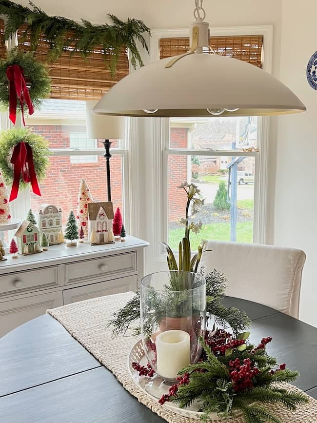 A cozy breakfast nook features a dark circular table adorned with greenery and a candle. The white pendant lamp provides lighting inspiration as it hangs above. Windows with greenery wreaths allow in daylight, while miniature holiday houses charm on a side table.