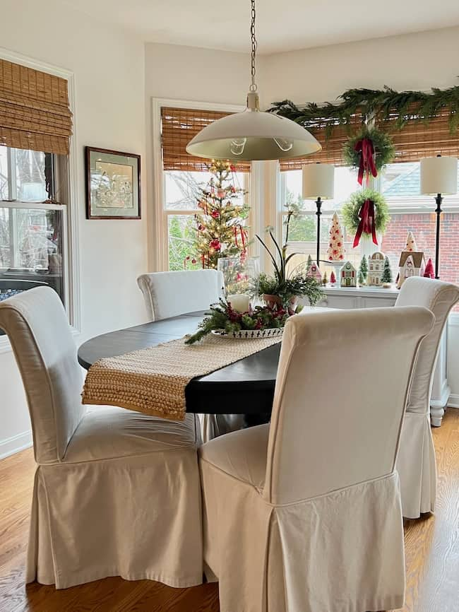 A cozy dining room with a round black table, surrounded by white slipcovered chairs, offers lighting inspiration. A festive centerpiece adorns the table while a Christmas tree, wreaths, and garlands enhance the holiday atmosphere. Natural light filters through the windows, illuminating this charming space.