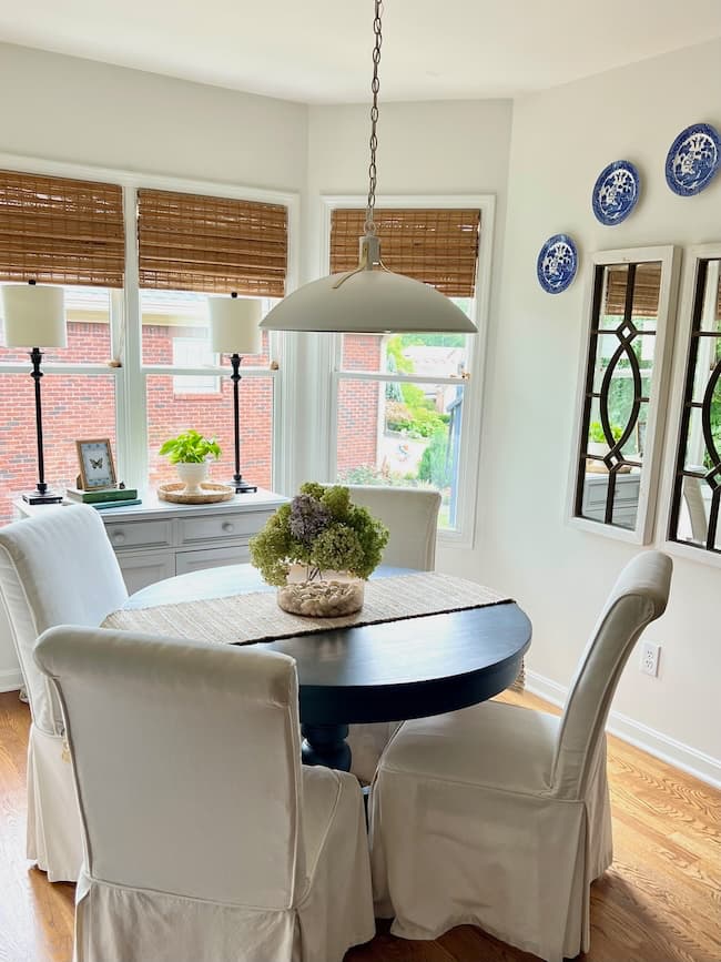 A cozy dining nook with a round black table, four white slipcovered chairs, breakfast nook lighting ideas including two lamps on a windowsill, bamboo blinds, a floral centerpiece, and decorative blue plates on a crisp white wall.