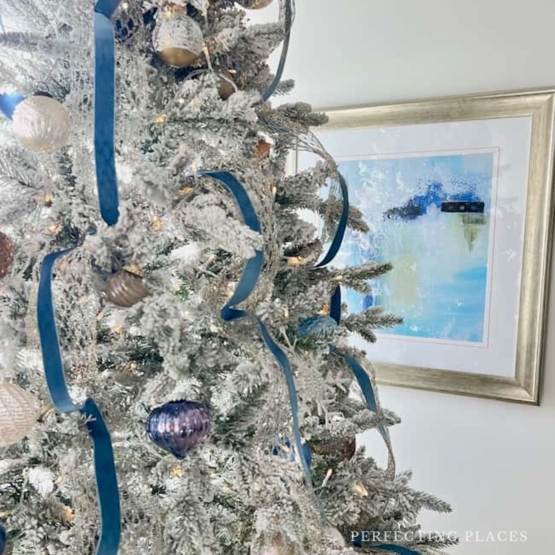 A cozy Christmas bedroom aesthetic features a flocked tree adorned with white lights, blue ribbons, and baubles. It stands elegantly in the room, complementing a framed abstract painting on the wall.