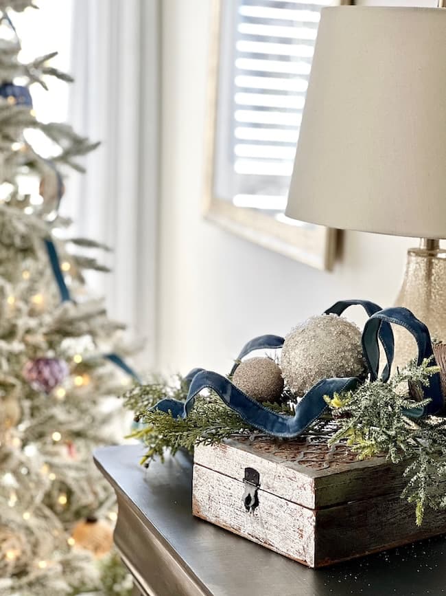 A festive holiday scene with a flocked Christmas tree in the background. In the foreground, a wooden box holds pine branches, blue ribbon, and ornaments, topped with a lamp. Soft light filters through window blinds, capturing the cozy Christmas bedroom aesthetic perfectly.