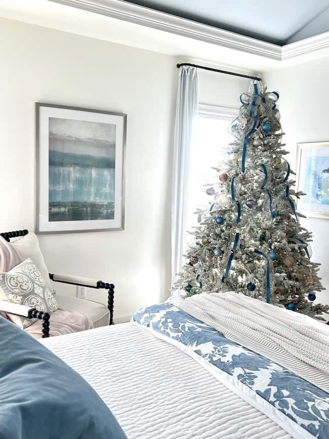 A cozy Christmas bedroom aesthetic unfolds with a stunning flocked tree adorned in blue and silver ornaments, ribbons, and lights. The room features a bed with blue floral bedding, an armchair with cushions, and a large abstract painting on the wall.