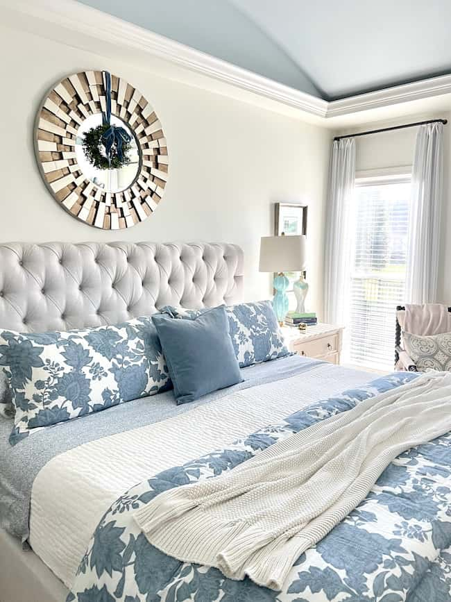 A cozy bedroom features a plush, tufted headboard and blue floral-patterned bedding. A wreath-adorned mirror hangs above, exuding a Christmas aesthetic. Beside the flocked tree, a bedside table with a lamp complements the window's sheer curtains in this serene blue decor.