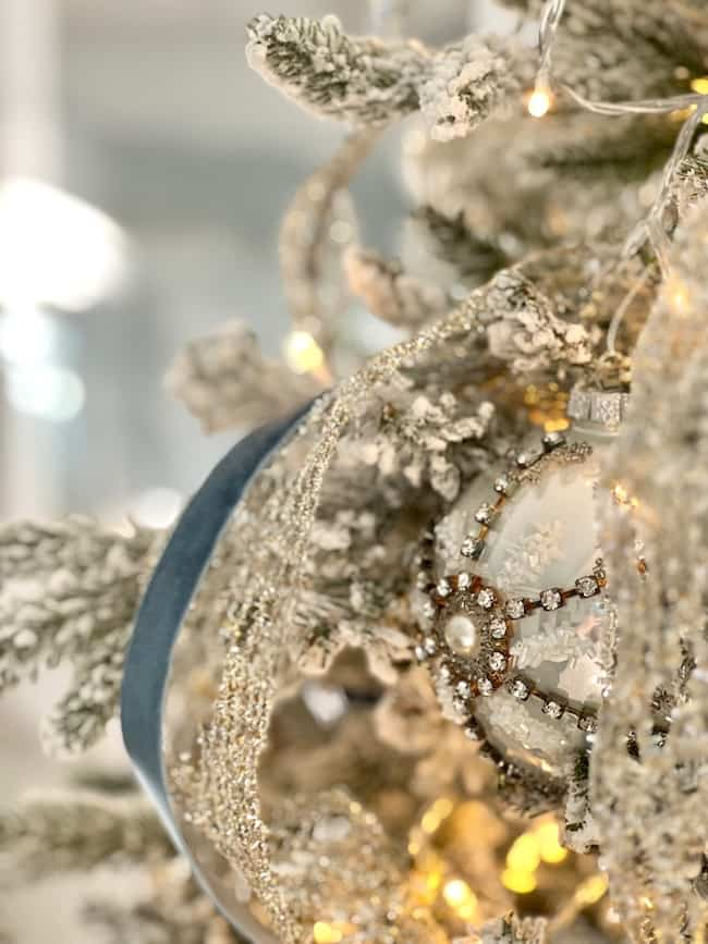 Close-up of a snow-dusted, flocked Christmas tree in a cozy bedroom aesthetic, adorned with a sparkling ornament featuring intricate gold and silver designs. Set against blue decor, the tree is draped with shimmering ribbon as twinkling lights illuminate its festive charm.