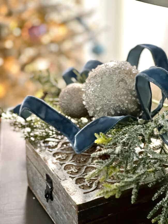 A decorative box topped with frosted ornaments, a blue ribbon, and greenery sits on a table, perfectly capturing the cozy Christmas bedroom aesthetic. In the background, a softly blurred flocked tree enhances the serene blue decor theme.