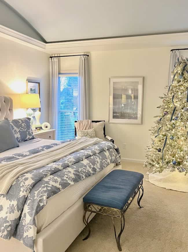 A cozy Christmas bedroom aesthetic unfolds with a flocked tree, blue and white floral bedding on a plush bed, and a bench at the foot. A nightstand with a lamp complements the beige armchair, as curtains frame large windows. Art adorns the walls, enhancing the serene blue decor.