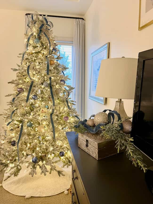 A beautifully decorated flocked Christmas tree with blue ribbons and ornaments stands in a warmly lit room. Nearby, a table capturing the cozy Christmas bedroom aesthetic displays a box filled with similar ornaments and a lamp, with curtains framing a window.