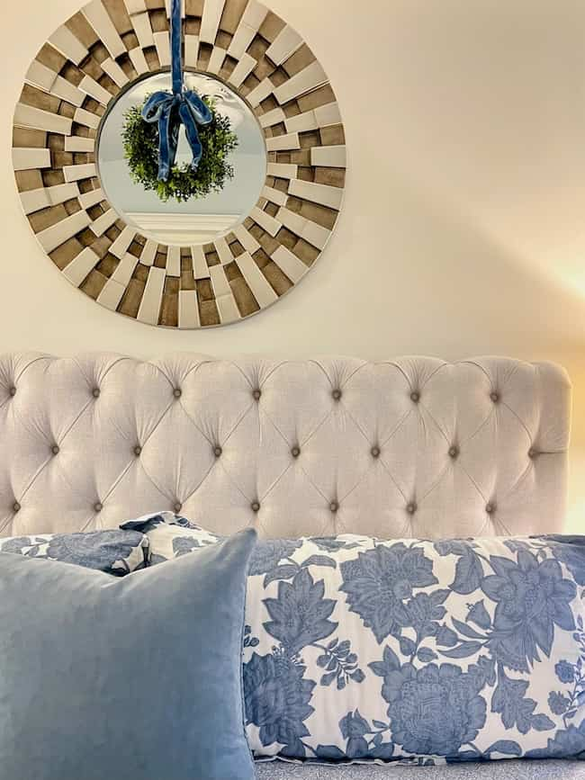 A cozy Christmas bedroom aesthetic features a tufted headboard and pillows with blue floral patterns. Above the bed hangs a round mirror with a wooden frame, adorned with a small wreath tied with a blue ribbon, complemented by a flocked tree for festive charm.