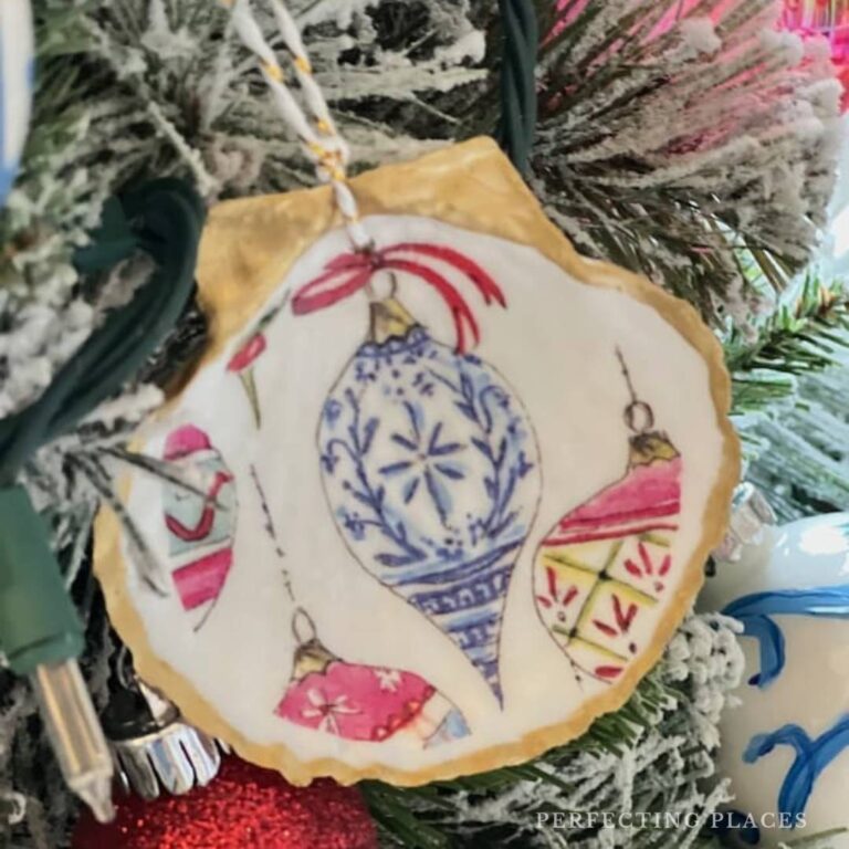 A festive Christmas ornament hanging on a frosted tree branch features an illustration of vintage-style baubles in blue, pink, and gold with intricate patterns, reminiscent of DIY decoupage seashell creations. The scene is accented by string lights and other decorations.