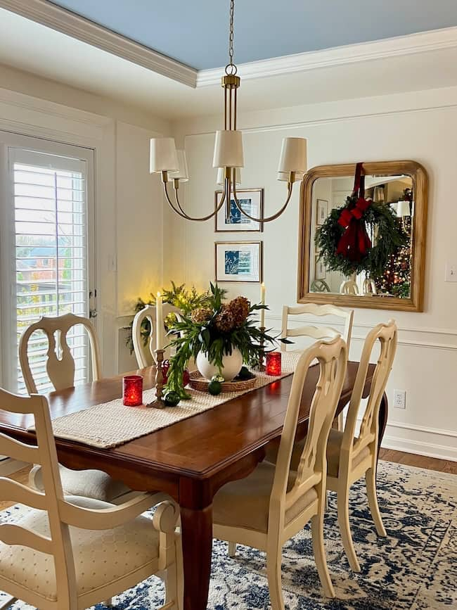 A dining room featuring a wooden table and six white chairs offers perfect lighting inspiration. The table is adorned with flowers, red candles, and a runner. Overhead, a chandelier glows warmly. A mirror with a wreath graces the wall, while natural light streams through shuttered doors.