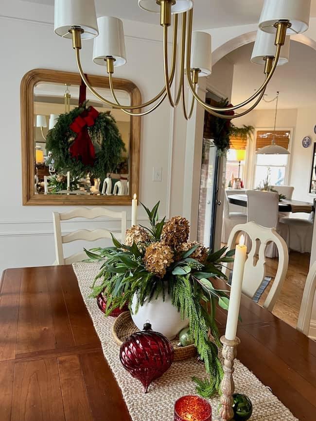 A dining table, beautifully illuminated by breakfast nook lighting, is decorated for the holidays. It features a centerpiece with green foliage and dried hydrangeas in a white vase. A burlap runner, candles, and red ornaments add warmth. A large wreath hangs on a mirror in the background.