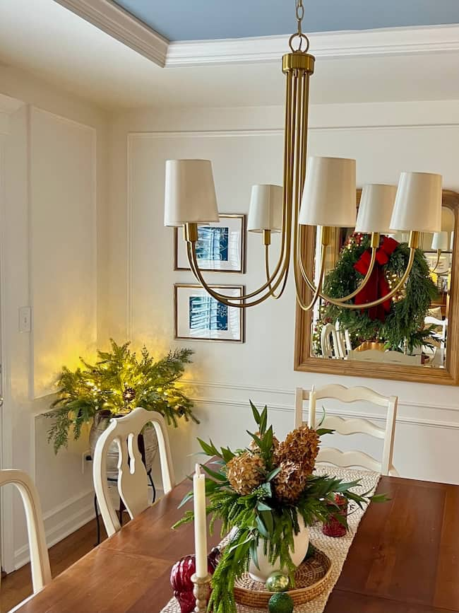 Elegant dining room with effortless style, featuring a wooden table adorned with floral centerpieces and candles. A gold chandelier provides exquisite dining room lighting. A mirror with a wreath graces one wall, and festive greenery decorates a corner, adding a warm holiday ambiance.