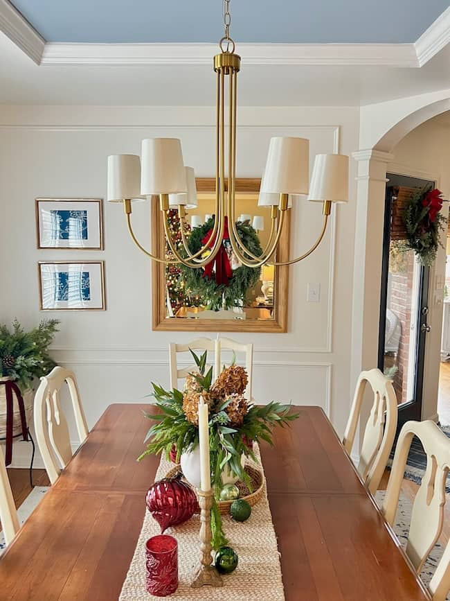 The dining room, a cozy breakfast nook, boasts a wooden table decorated with a festive centerpiece featuring green foliage and red ornaments. A gold chandelier with white lampshades provides lighting inspiration. The space is framed by white walls, framed art, and a glass door adorned with a wreath.