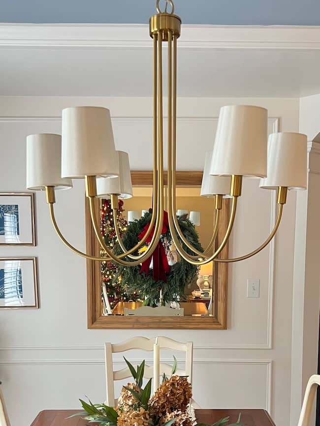 A modern gold chandelier with six white lampshades provides lighting inspiration over a dining table. In the background, a decorative wreath with a red ribbon is reflected in a mirror. A chair and framed pictures add charm to this elegant dining room.