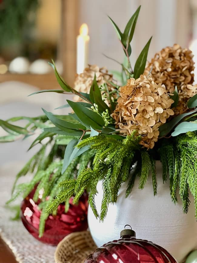 A festive centerpiece graces the dining room table, with dried hydrangeas, lush greenery, and eucalyptus in a white vase. Red ornaments and a lit candle provide lighting inspiration for the holiday decor on a textured tablecloth.