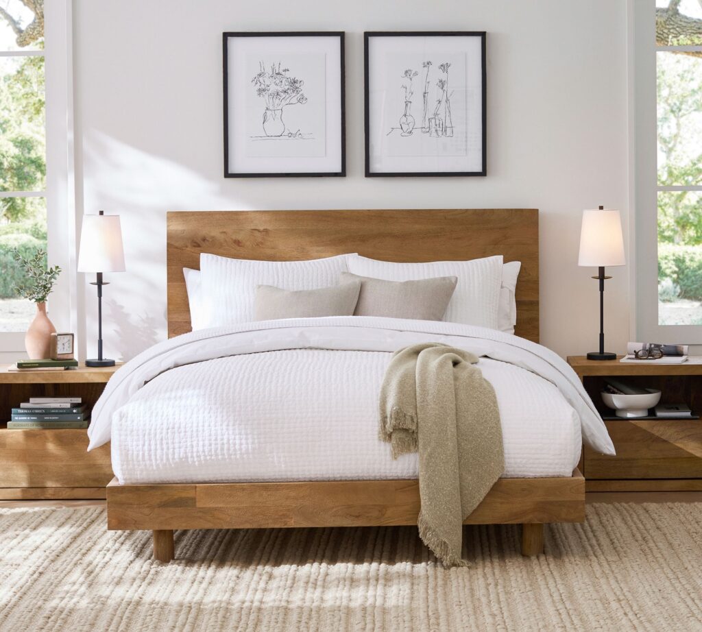 A cozy bedroom with a wooden bed frame and white bedding, featuring two beige pillows. Two minimalist framed art pieces hang above the bed. Beside a flocked tree, the festive decor is accented with blue touches. Bedside tables with matching lamps sit by large windows, and a beige throw drapes over the bed.