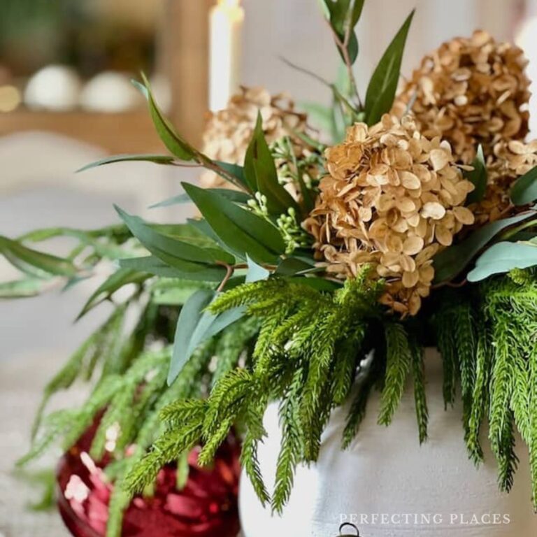 A festive floral arrangement with dried hydrangeas, lush evergreen branches, and green leaves in a sleek white vase. A red, glossy ornament peeks out beside it, creating a Seven on Saturday holiday ambiance.