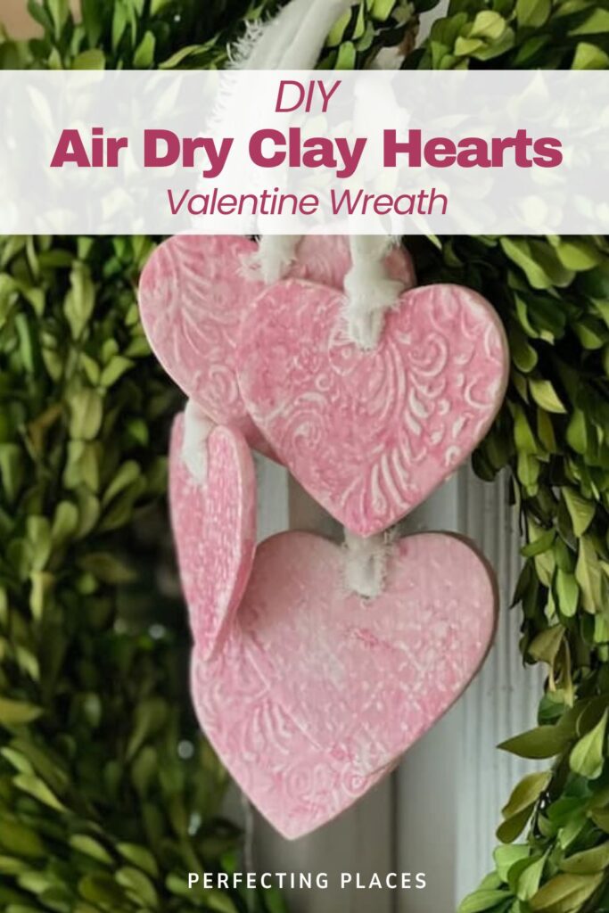 A close-up of three pink heart-shaped ornaments with embossed designs hanging gracefully from a lush greenery wreath. The text reads "DIY Air Dry Clay Hearts Valentine’s Day Wreath" and "Perfecting Places" at the bottom.