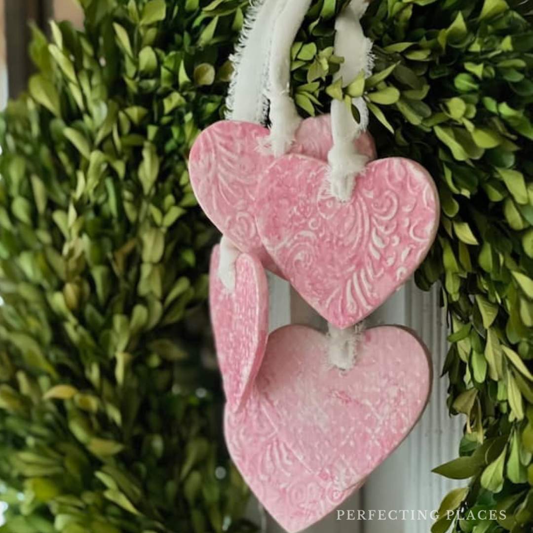 Three pink heart-shaped ornaments with intricate patterns hang on a vibrant green Valentine's Day wreath. The clay hearts are delicately attached by white strings, adding a touch of charm and elegance.
