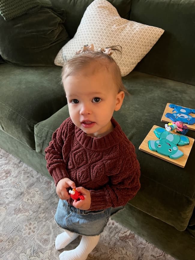 A toddler with light brown hair stands in front of a green couch, bringing a touch of Spring Refresh to the room. Wearing a maroon sweater and denim pants, the child holds red toy figures while colorful animal puzzle pieces rest on a soft rug nearby.