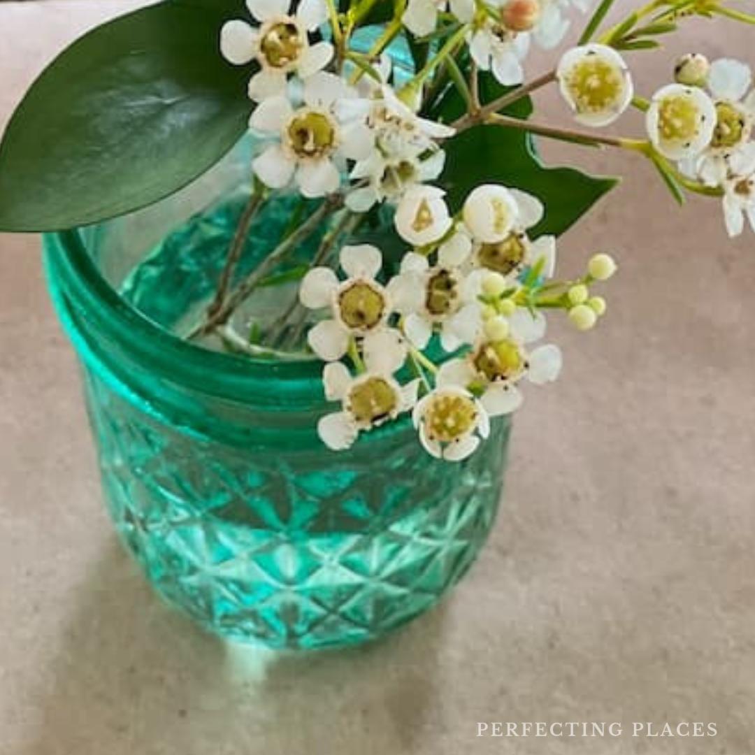 A turquoise glass jar with a diamond pattern cradles a bouquet of small white flowers and green leaves, capturing an essence of serenity. Resting on a beige surface, it hints at the comforts of "Things I'm Loving," while "Perfecting Places" graces the bottom right corner.